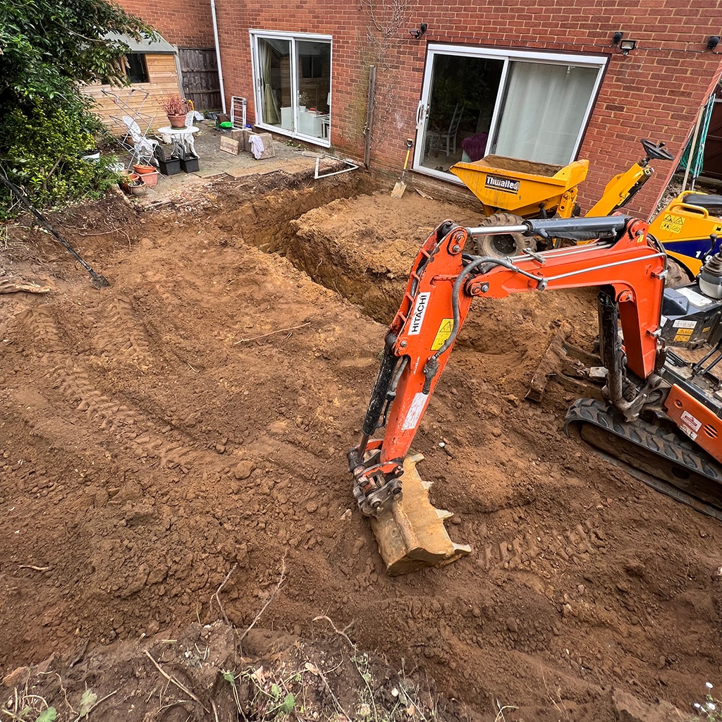 Mini digger and Thwaites tracked dumper positioned at an excavated footings trench during a man and digger groundwork job in a residential back garden.