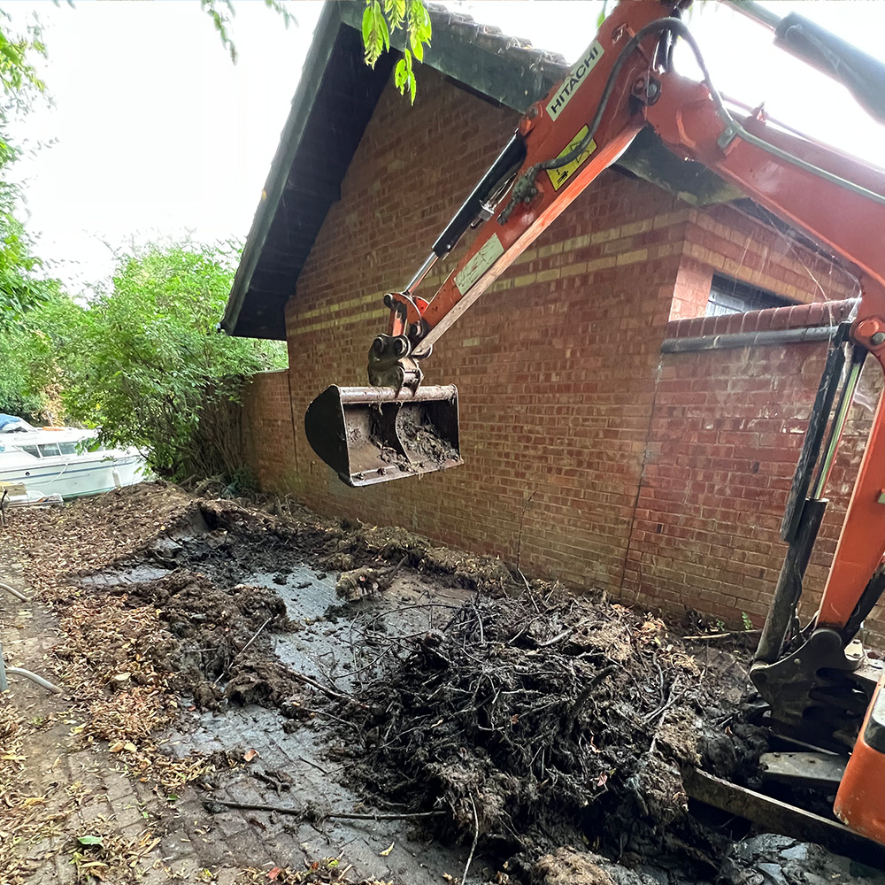 Mini digger clearing spoil and tree roots tight against a house wall on a Newport Pagnell man and digger groundwork job.
